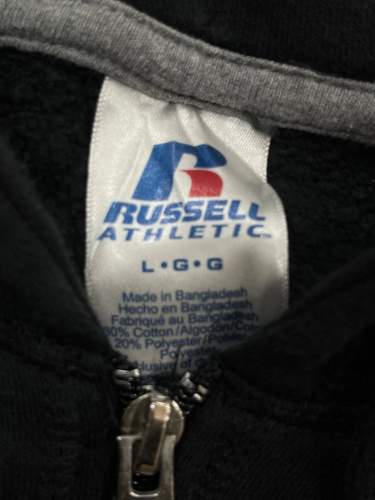 Close-up of a Russell Athletic L-G size garment tag sewn inside a black zippered jacket, showing the brand logo and fabric composition details.