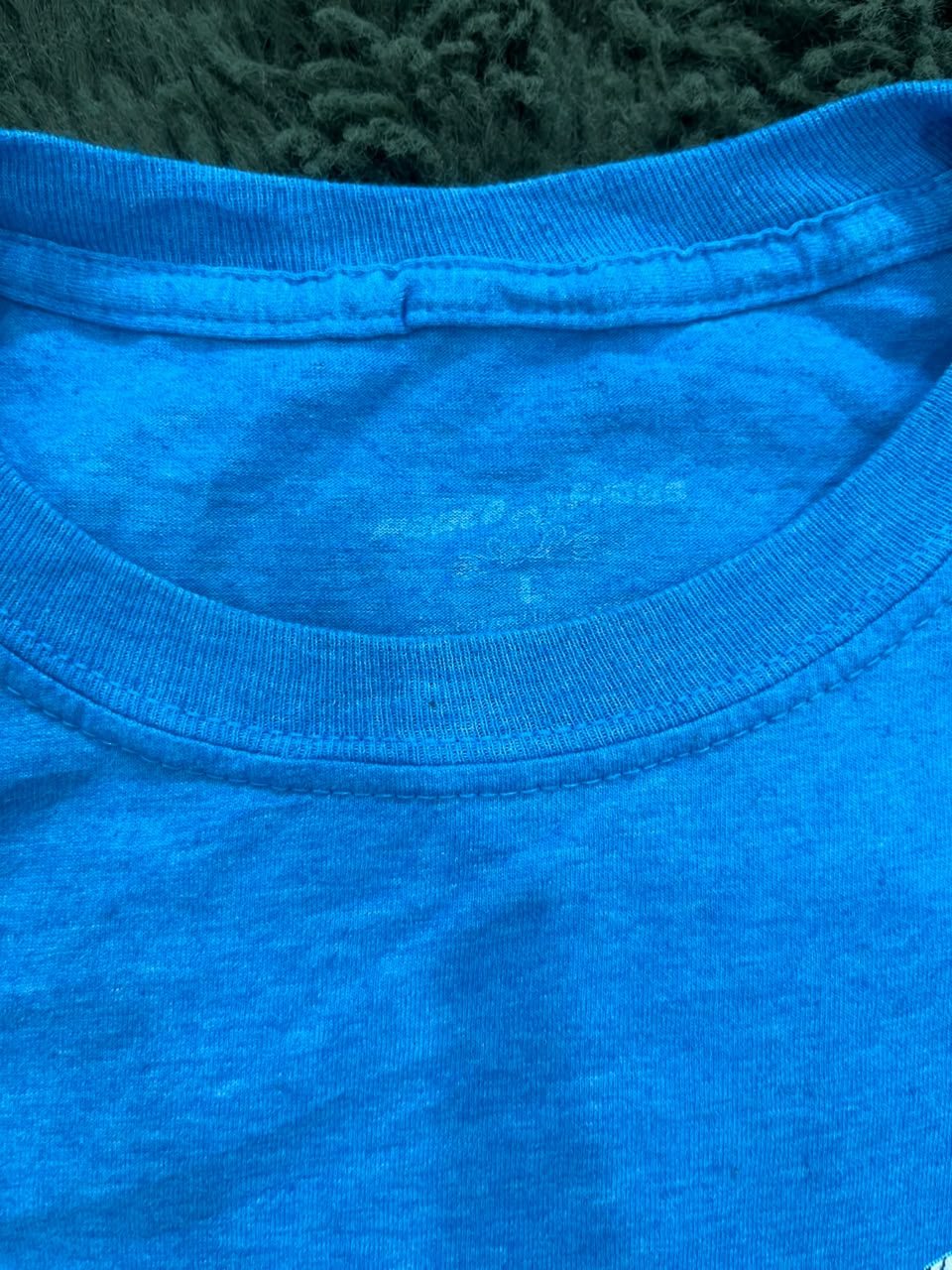Close-up of a bright blue crewneck t-shirt showing the inner neck label and ribbed collar, with a dark textured background visible at the top.