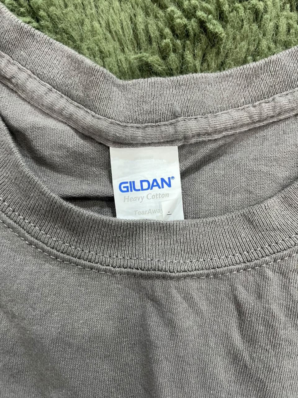 Close-up of a gray Gildan Heavy Cotton t-shirt neck label, showing the brand name in blue text on a white tag, with a green textured fabric background.