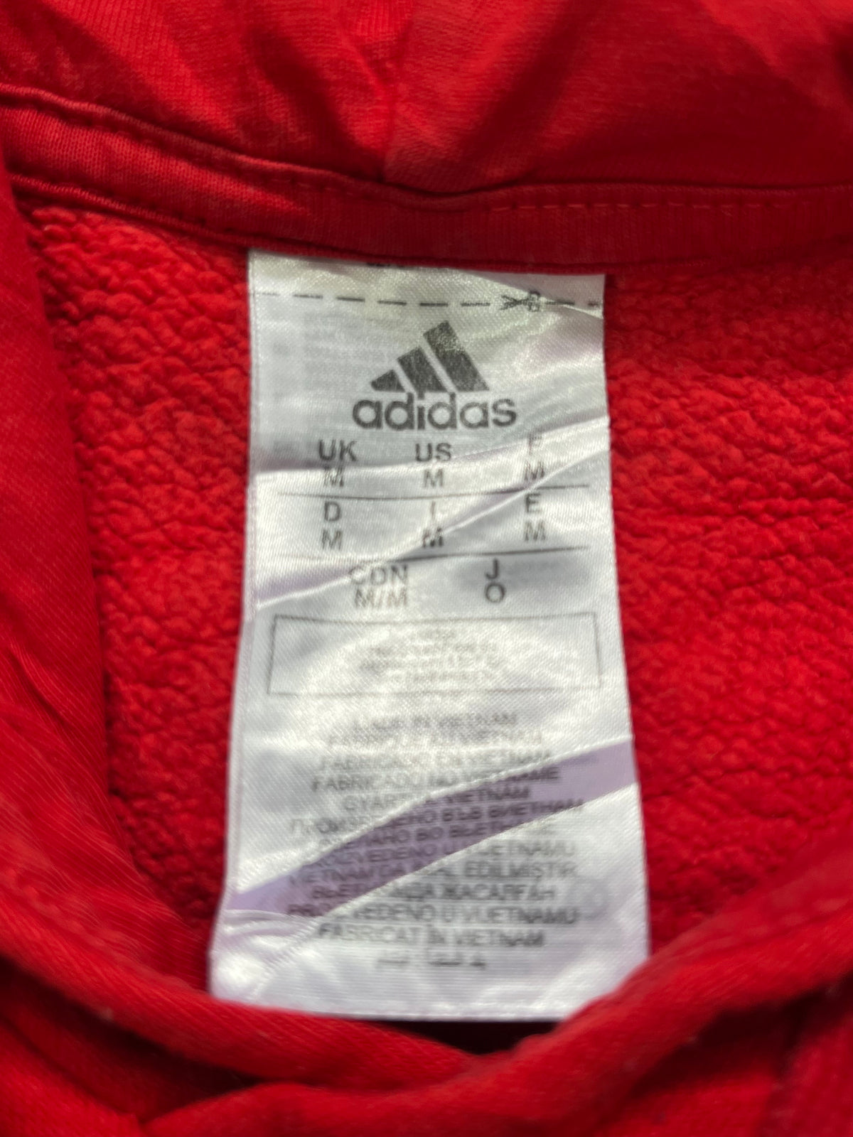 Close-up of an Adidas garment interior label sewn onto a bright red fabric background, showing the Adidas logo and size information.