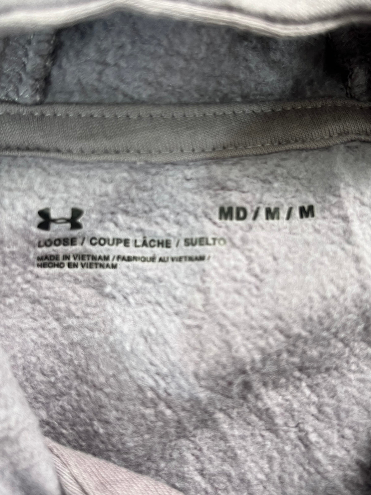 Close-up of the inside neck label of a gray Under Armour fleece garment, showing the Under Armour logo, 'LOOSE / COUPE LÀCHE / SUELTO' text, and 'MD / M / M' size marking.