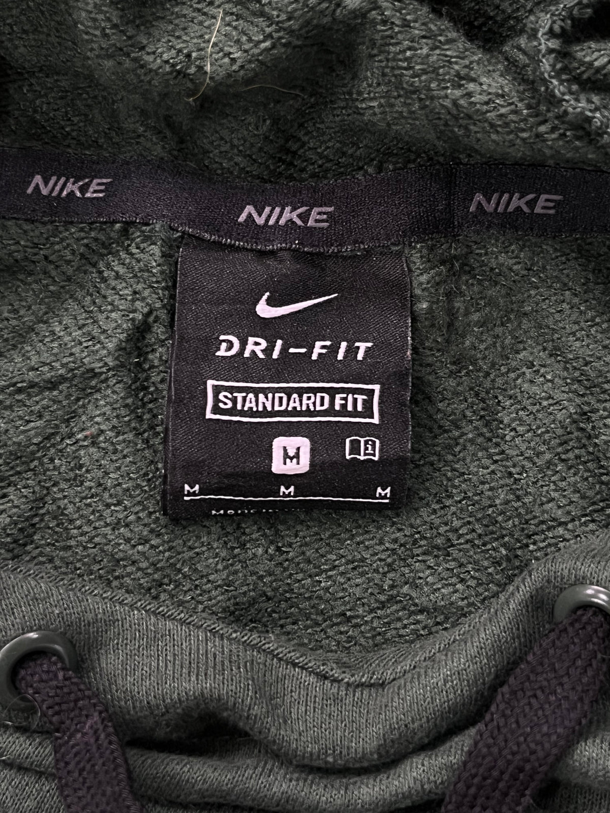 Close-up of a Nike Dri-Fit Standard Fit hoodie label on a dark olive green textured fabric, with drawstrings visible at the bottom corners.