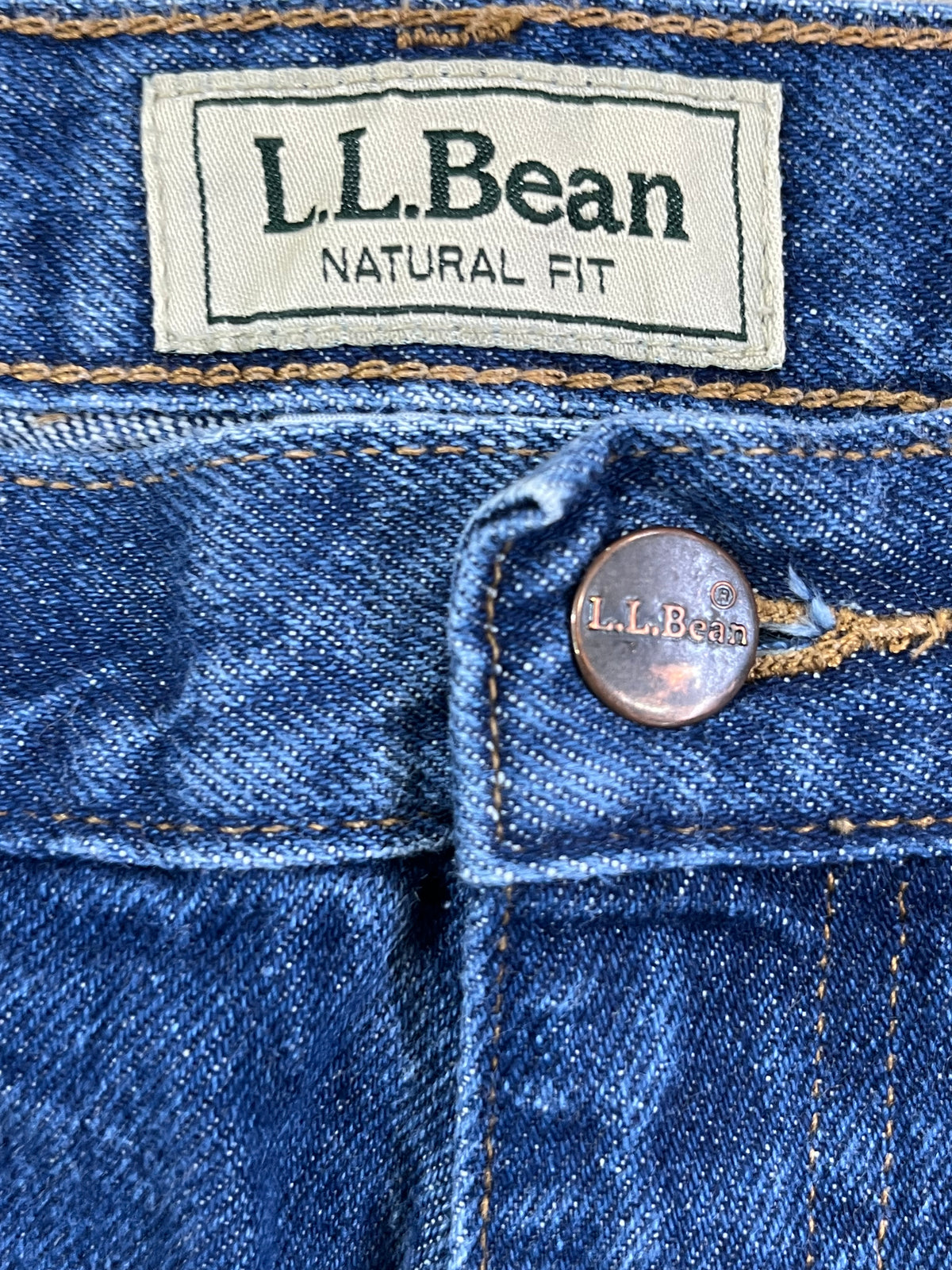 Close-up of blue denim jeans showing the waistband, a cream-colored L.L.Bean brand label, and a copper-toned metal button.