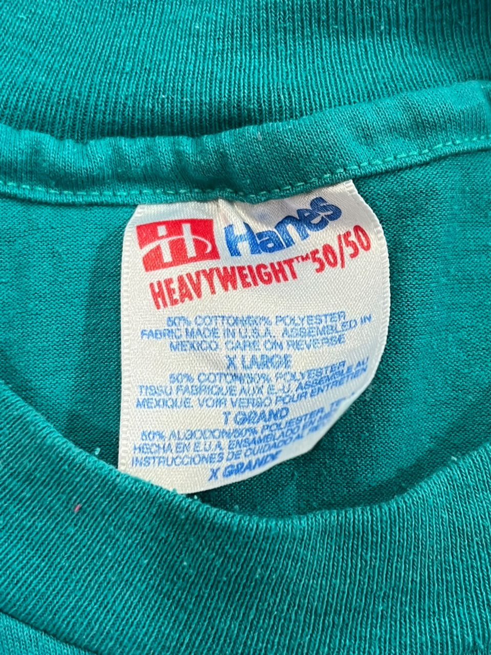 Close-up of a teal green garment neck label reading Hanes Heavyweight 50/50, with the Hanes logo in red and blue on a white tag.