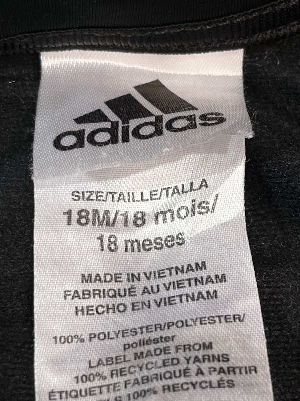 Close-up of an Adidas garment size label showing the brand logo, size 18M/18 months, and material details on a black garment.