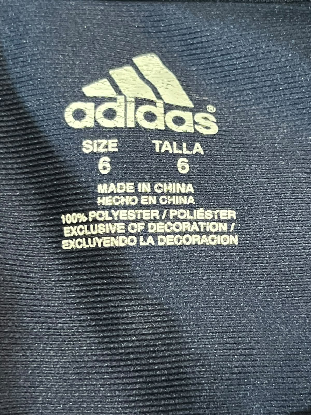 Close-up of an Adidas garment label printed in light beige on a dark navy ribbed fabric background.
