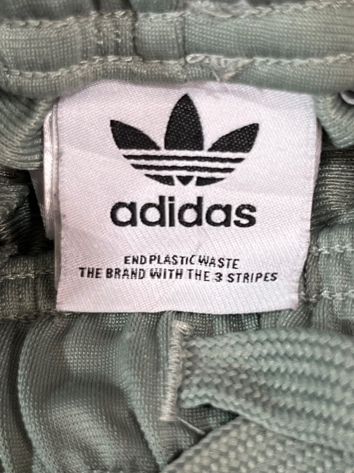 Close-up of an Adidas brand label sewn onto sage green fabric, showing the black trefoil logo and text 'adidas' and 'END PLASTIC WASTE THE BRAND WITH THE 3 STRIPES'.