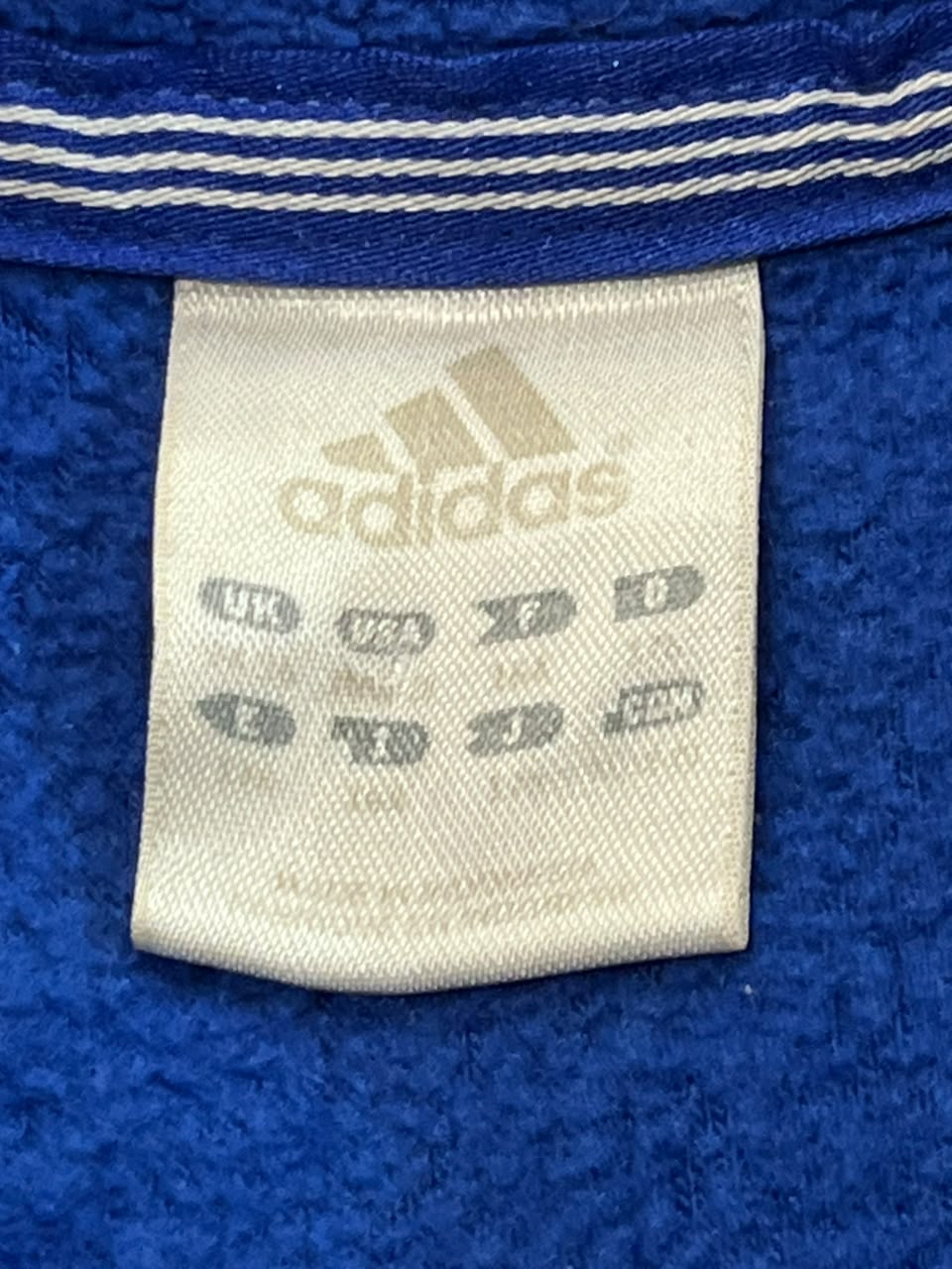 Close-up of an Adidas brand tag sewn onto a textured royal blue fabric, with a matching blue and white striped trim visible at the top.