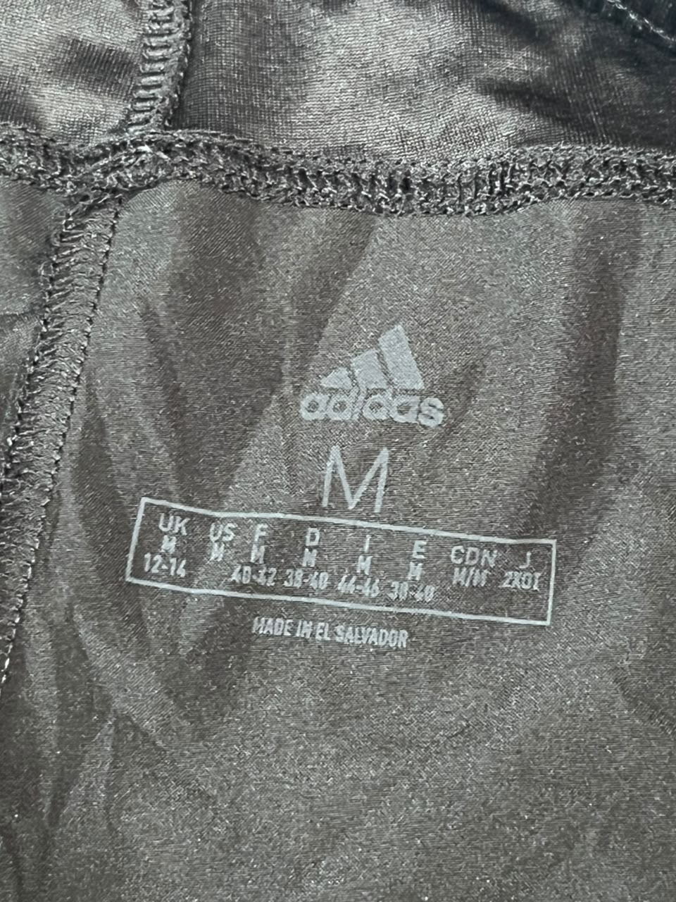 Close-up of the inside neck label of a dark gray Adidas garment, showing the Adidas logo, size M, and size conversion chart.