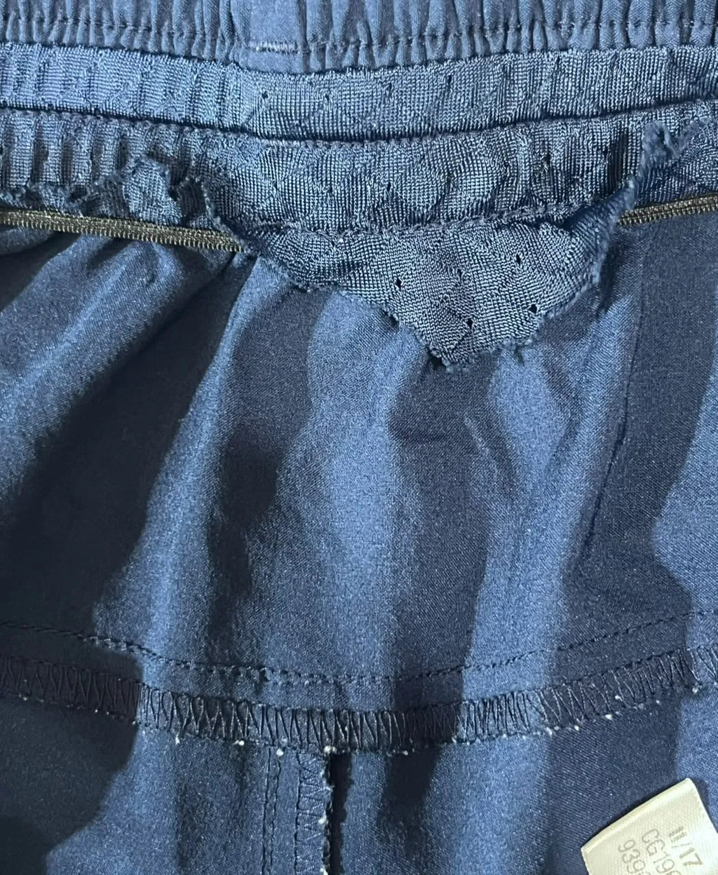 Close-up of a blue-gray fabric garment showing the waistband, stitching, and a small white size tag in the lower right corner.
