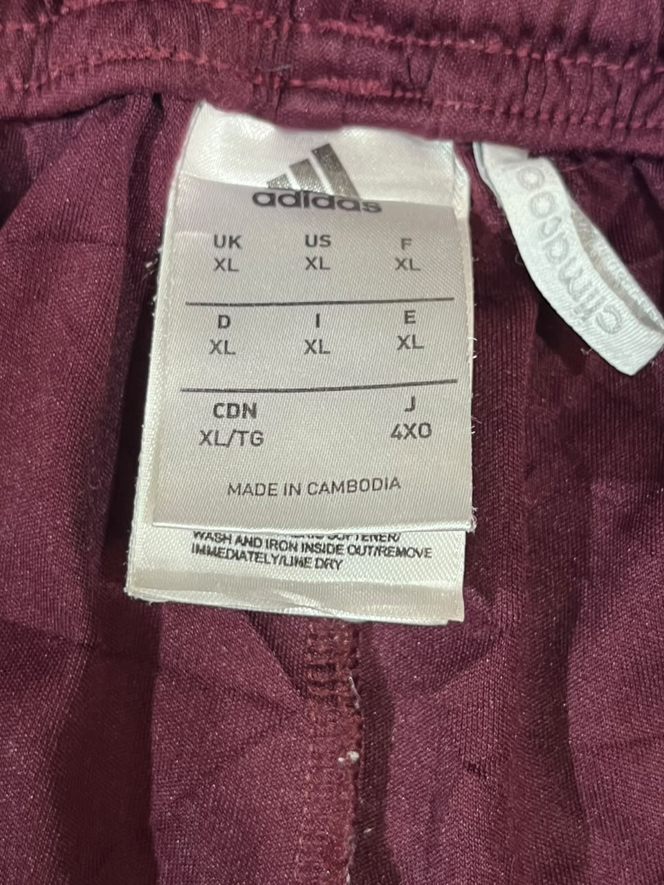 Close-up of a burgundy Adidas garment label showing size information for UK, US, and CDN sizes, with the Adidas logo visible on the white tag.