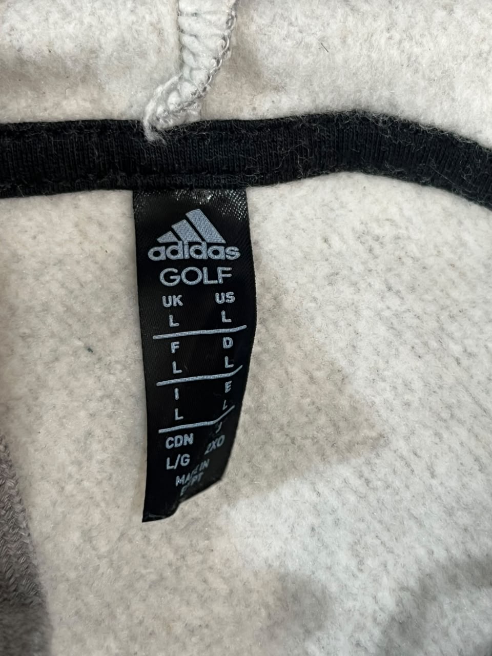 Close-up of an Adidas Golf garment label sewn onto a light gray heathered fabric, with a black elastic band and white drawstring visible.