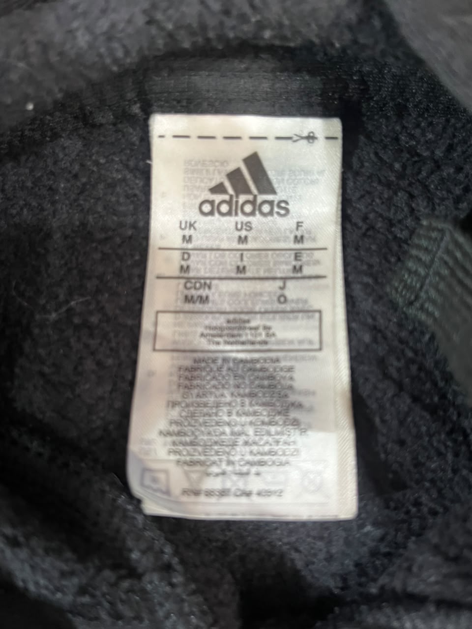 A close-up of an Adidas garment size and care label sewn onto the inside of a black textured fabric garment.