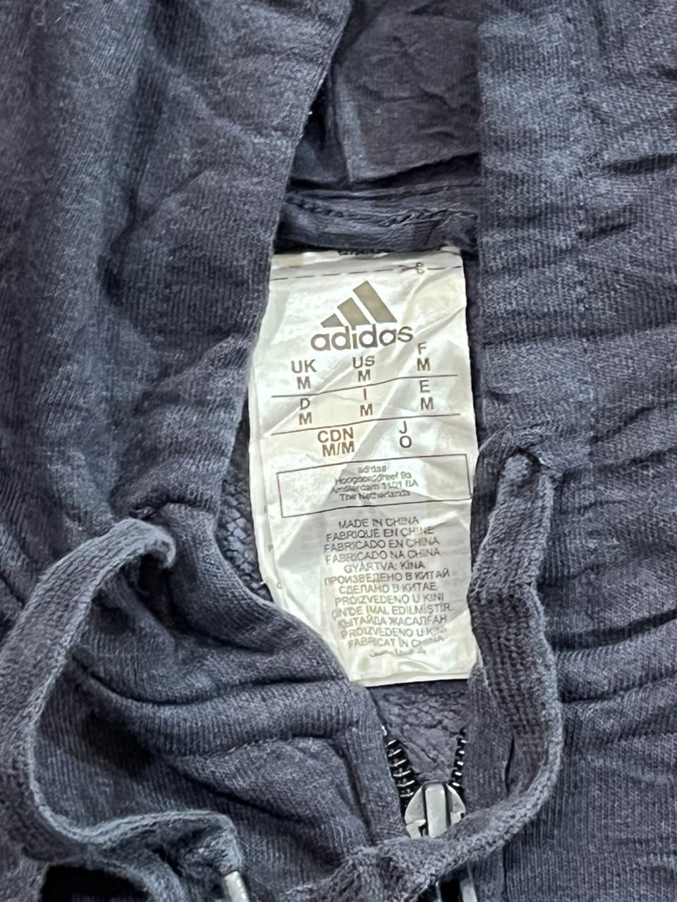 Close-up of an Adidas garment label sewn into the inside neckline of a dark gray ribbed knit hoodie or sweatshirt, with drawstrings visible at the front.