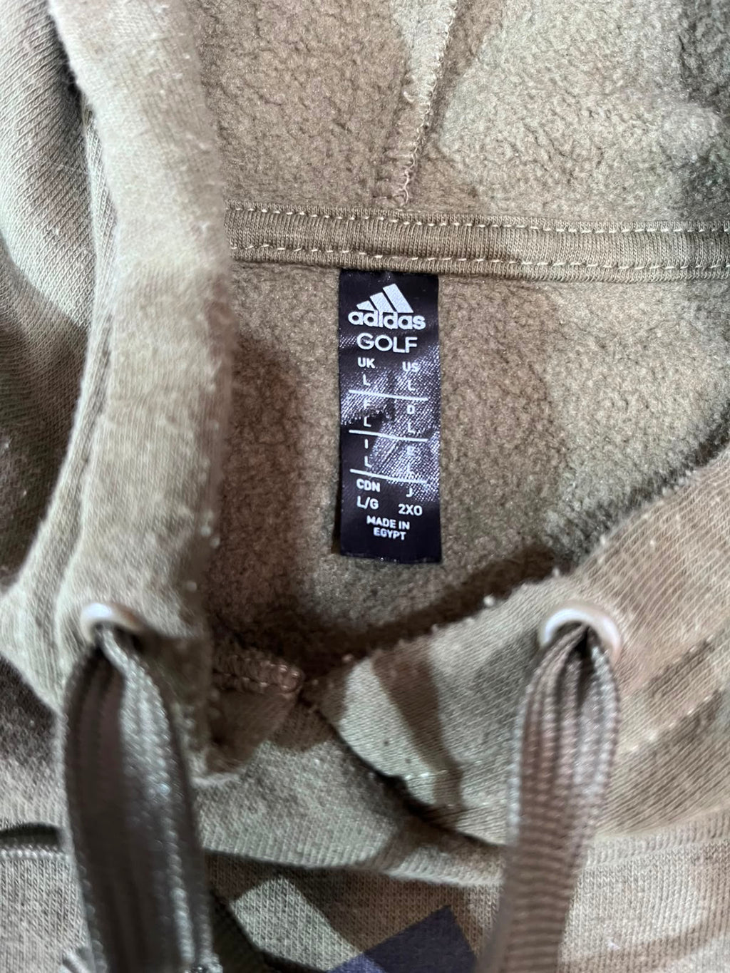 Close-up of the inside neck of a beige Adidas Golf hoodie, showing the black size label with measurements and drawstrings at the bottom.
