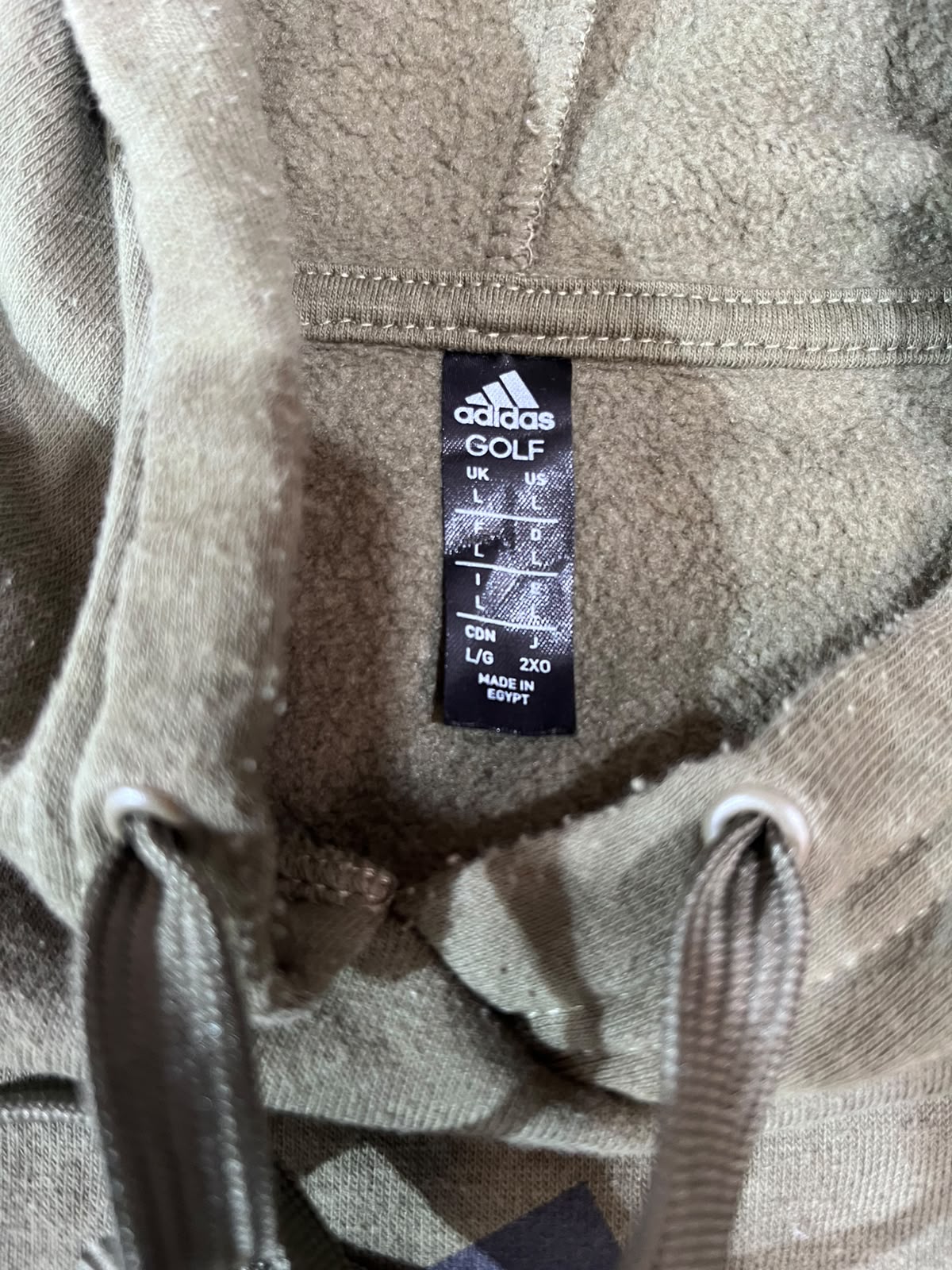 Close-up of the inside neck of a beige Adidas Golf hoodie, showing the black size label with measurements and drawstrings at the bottom.