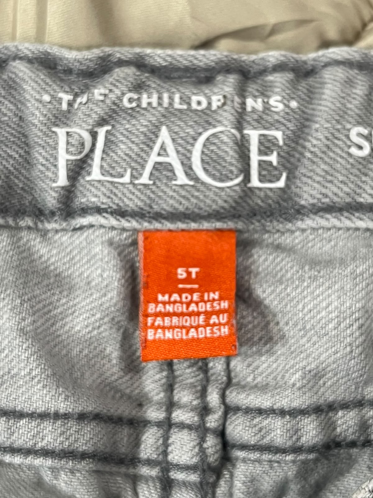 Close-up of a gray denim garment waistband with white printed text reading 'THE CHILD PINS PLACE' and an orange size/origin label that says '5T MADE IN BANGLADESH'.