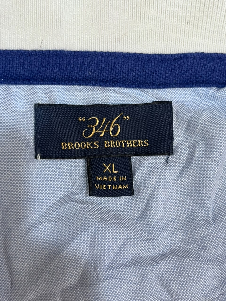 Close-up of a Brooks Brothers garment label showing the brand name "346" in gold on a black tag, with size XL and Made in Vietnam below, set against a light blue-gray textured fabric.