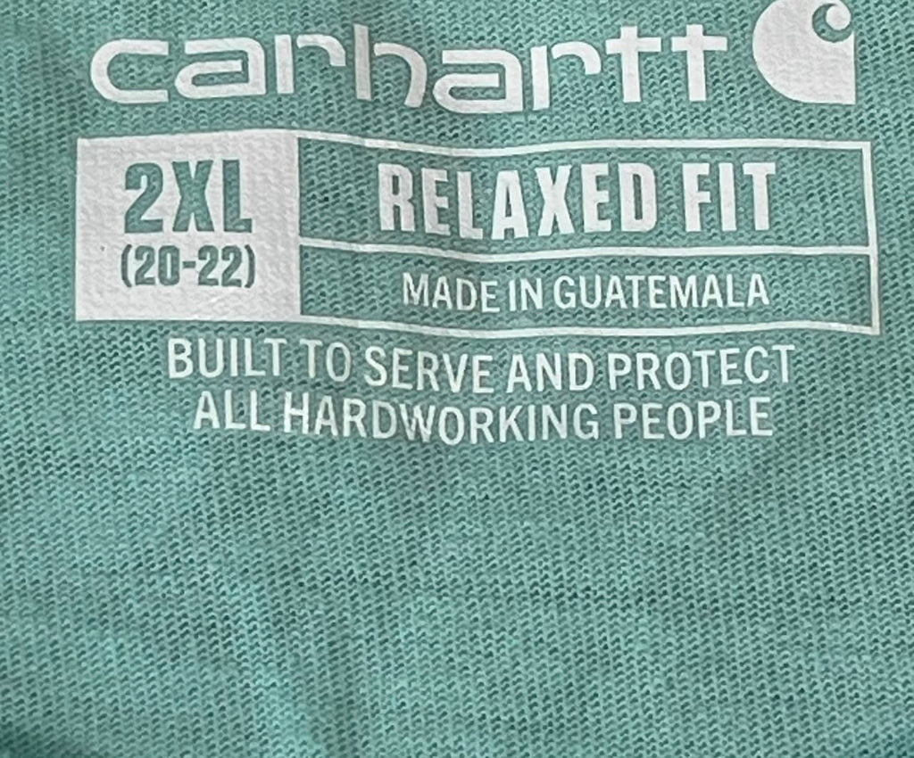 Close-up of a teal-green Carhartt fabric with white printed text reading '2XL RELAXED FIT', 'MADE IN GUATEMALA', and 'BUILT TO SERVE AND PROTECT ALL HARDWORKING PEOPLE'.