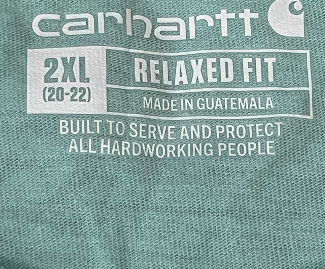 Close-up of a teal-green Carhartt fabric with white printed text reading '2XL RELAXED FIT', 'MADE IN GUATEMALA', and 'BUILT TO SERVE AND PROTECT ALL HARDWORKING PEOPLE'.