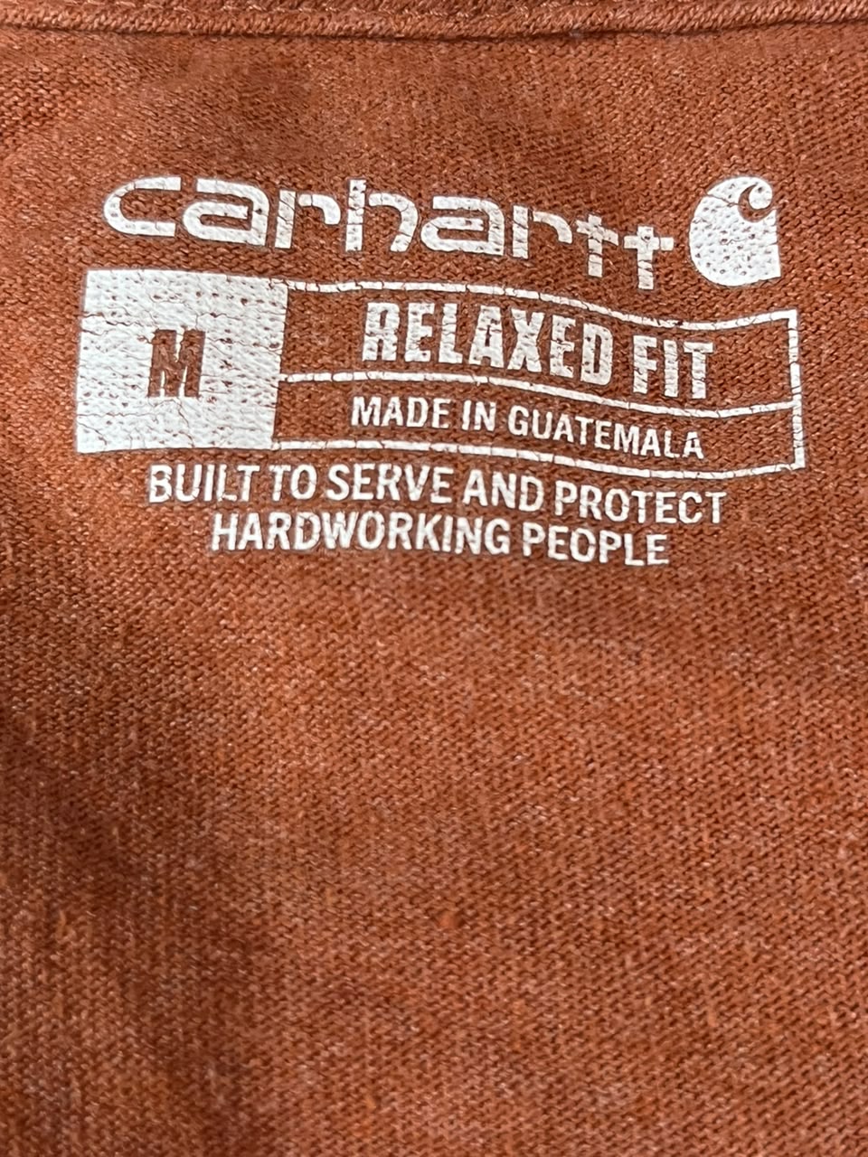Close-up of a brown Carhartt fabric label printed with white text reading 'carhartt', 'RELAXED FIT', 'MADE IN GUATEMALA', and 'BUILT TO SERVE AND PROTECT HARDWORKING PEOPLE'.