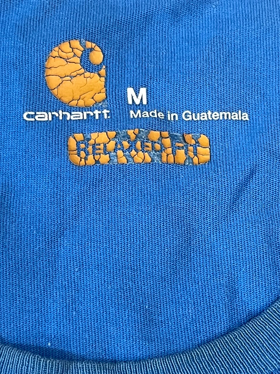 Close-up of a blue Carhartt garment with orange and white printed branding, including the Carhartt logo, size M, 'Made in Guatemala,' and a distressed orange 'GREAT' graphic.