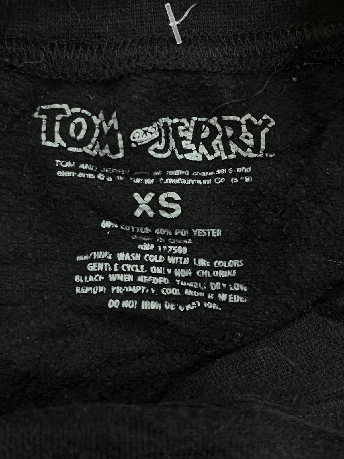 Close-up of a black garment's interior neck label area showing white printed text including 'Tom Jerry', 'XS', and care instructions.