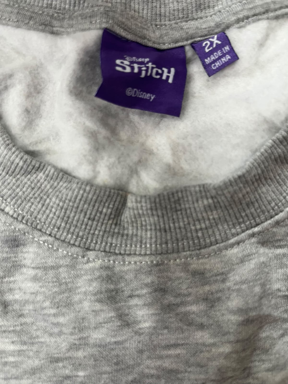 Close-up of a gray heathered sweatshirt or sweater neck with a white sherpa lining and visible sewn-in labels. One label reads 'Stitch' and the other says 'Made in China 2X'.