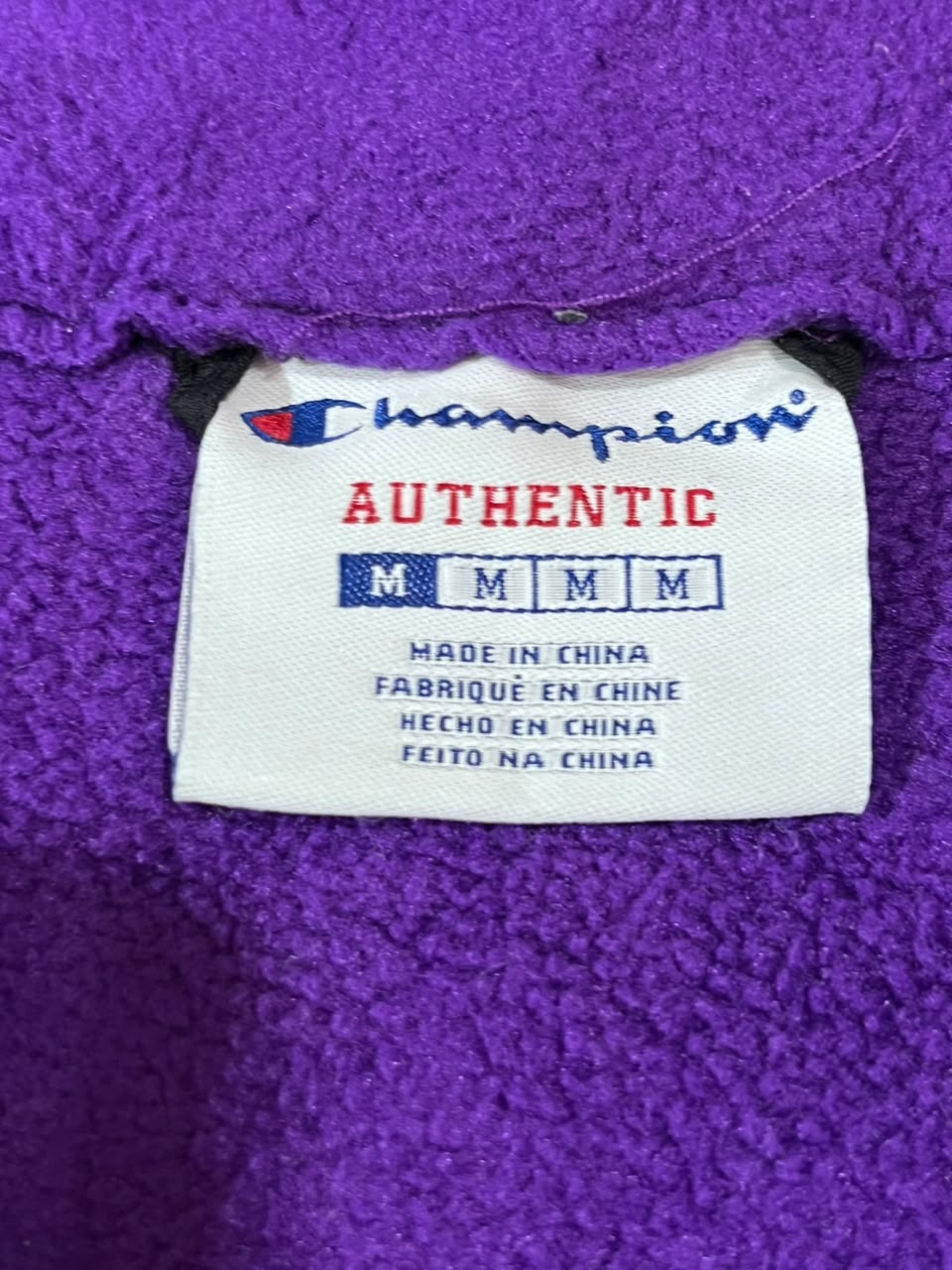 A close-up of a purple textured fleece garment showing a white Champion Authentic label sewn inside the collar.