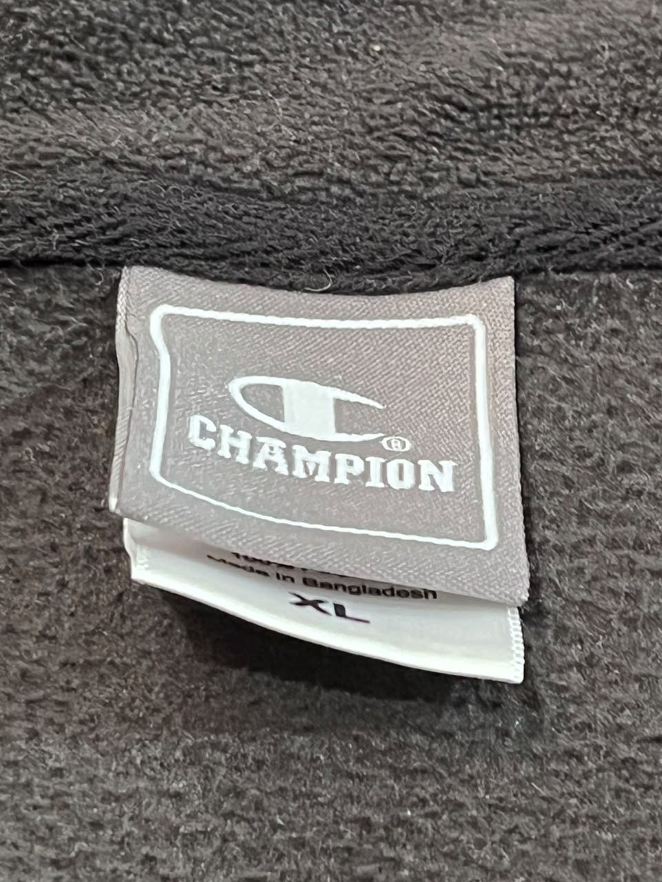 Close-up of a Champion brand fabric tag sewn onto a dark gray textured garment, with a white size tag below showing 'XL'.
