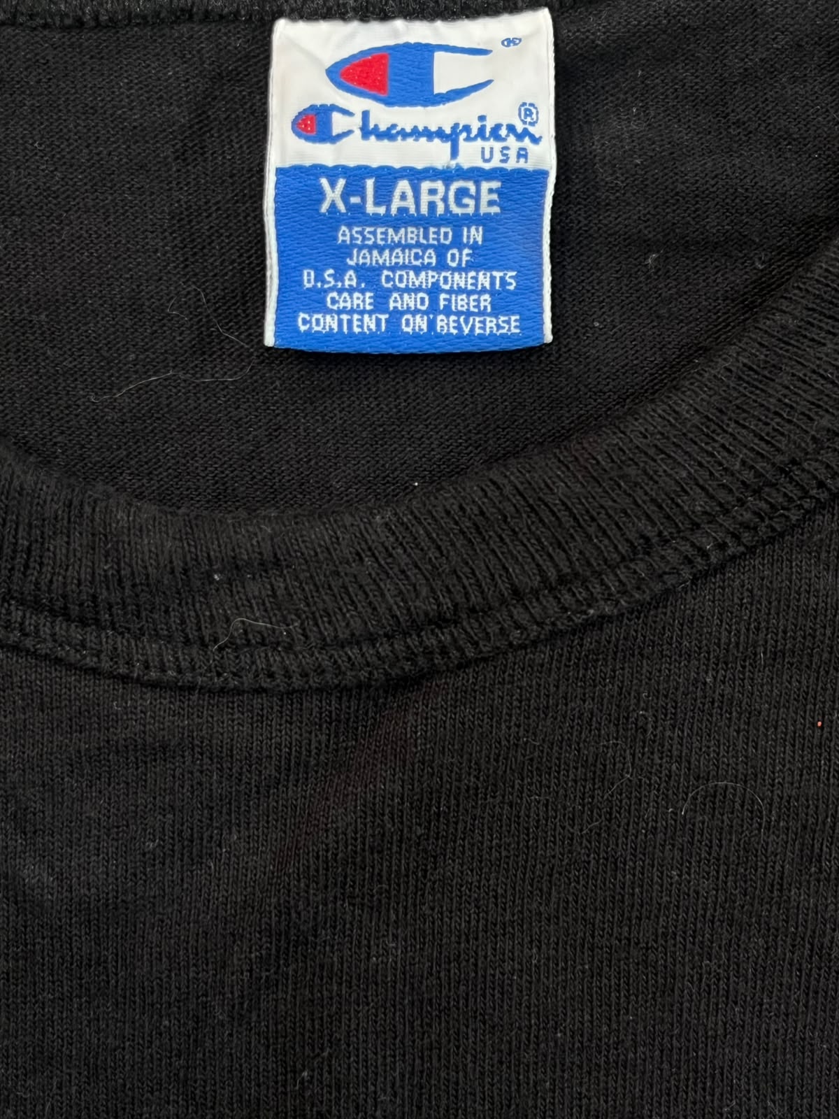 Close-up of a black crewneck sweatshirt showing the inside neck label which is white and blue with the Champion USA logo and text reading 'X-LARGE'.