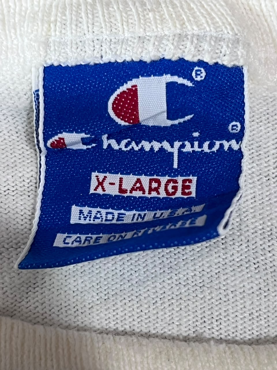 Close-up of a blue Champion knit garment label sewn onto a white ribbed fabric, showing the Champion logo and text reading X-LARGE, MADE IN U.S.