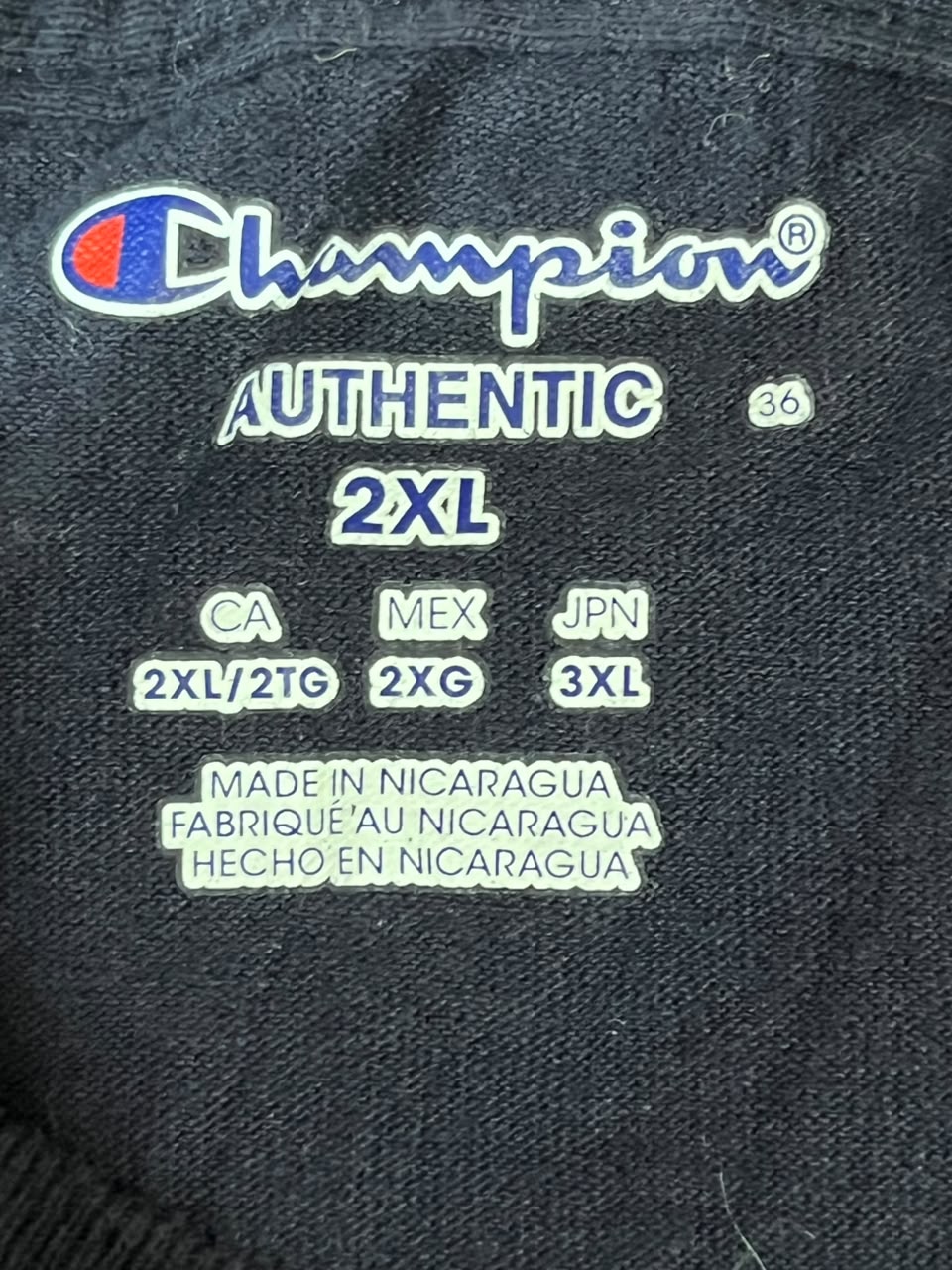 Close-up of a Champion Authentic 2XL garment label sewn onto dark charcoal fabric, showing the Champion logo, size information, and 'Made in Nicaragua' text.