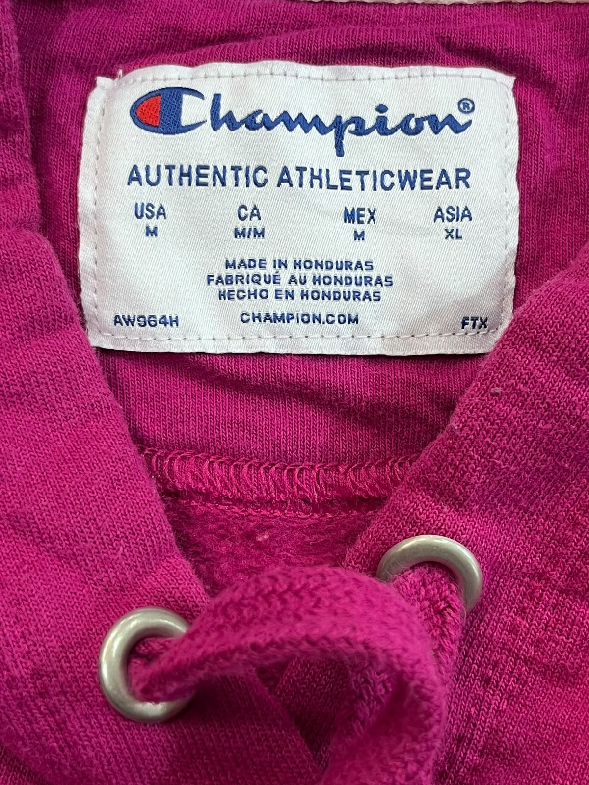 Close-up of a Champion athletic hoodie neck label, showing the brand name and size information, with bright pink fabric and metal drawstring rings visible.