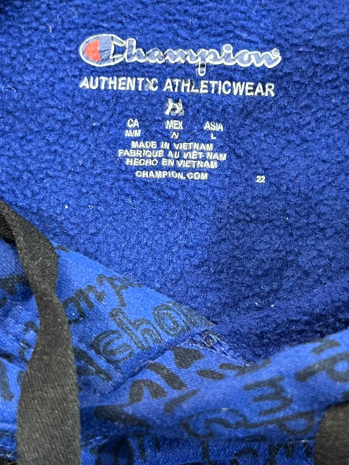 Close-up of a blue Champion athletic towel with white embroidered Champion branding and size information, with part of a black patterned fabric visible in the lower left corner.