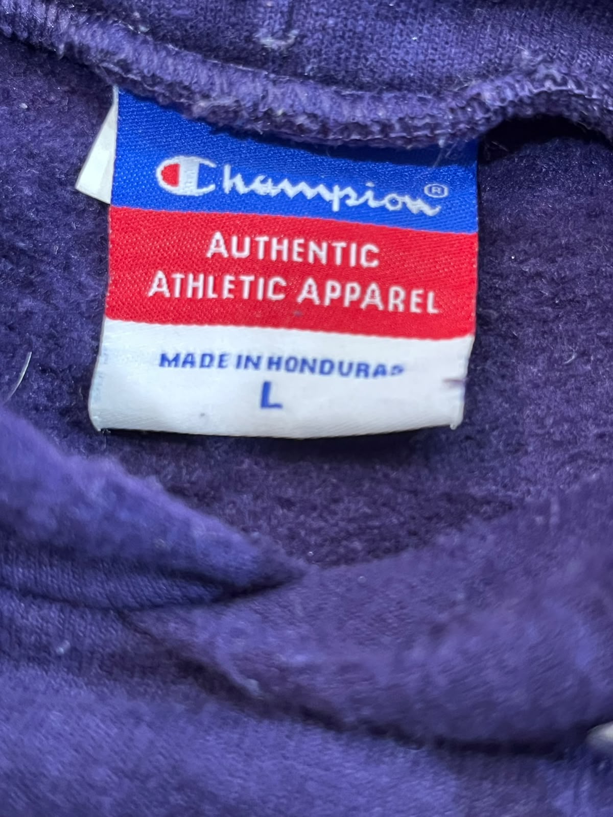 Close-up of a Champion Authentic Athletic Apparel size L garment label sewn onto purple fabric.