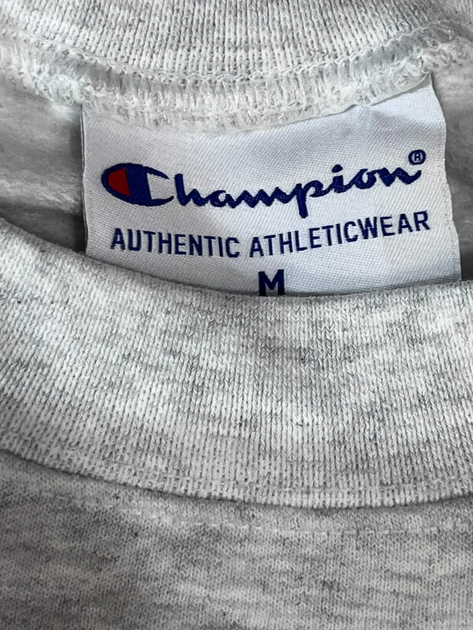 Close-up of a light gray heather crewneck sweatshirt showing the inside neck label with the Champion logo and 'AUTHENTIC ATHLETICWEAR' text.