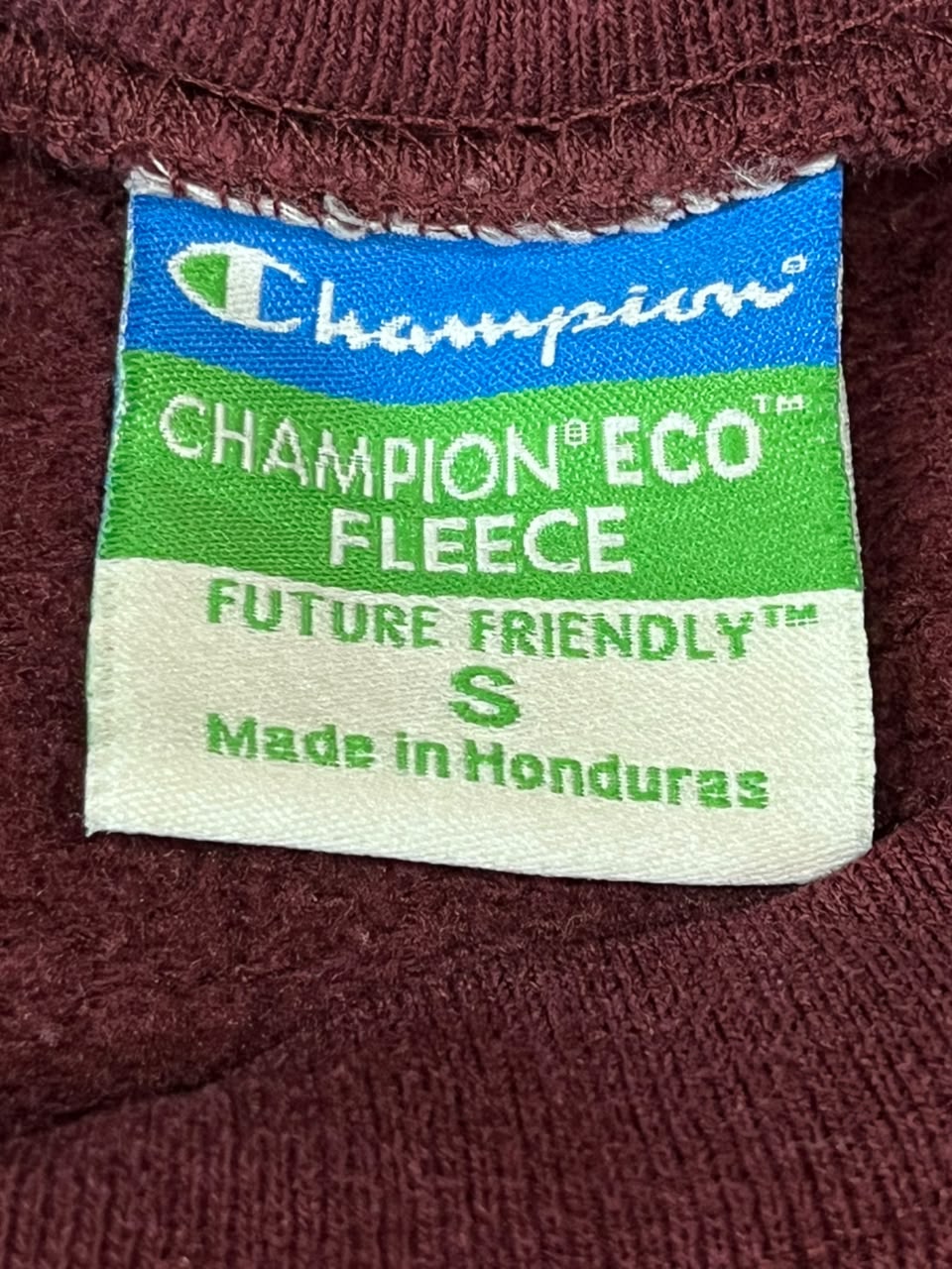 Close-up of a Champion Eco Fleece size S garment label sewn onto a dark burgundy knit fabric.