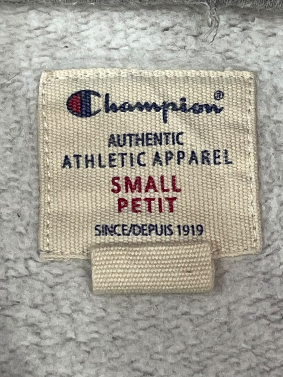 A close-up of a Champion brand fabric tag sewn onto a light gray textured garment. The tag is beige with the Champion logo in navy and red, and the text 'AUTHENTIC ATHLETIC APPAREL', 'SMALL', and 'SINCE/DEPUIS 1919' in navy and red.