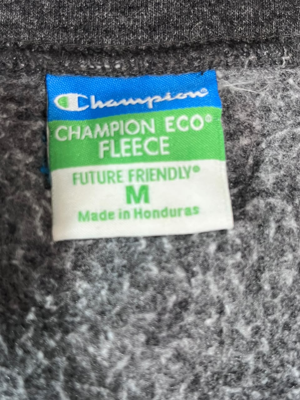Close-up of a Champion Eco Fleece garment tag on a gray textured fleece fabric background.
