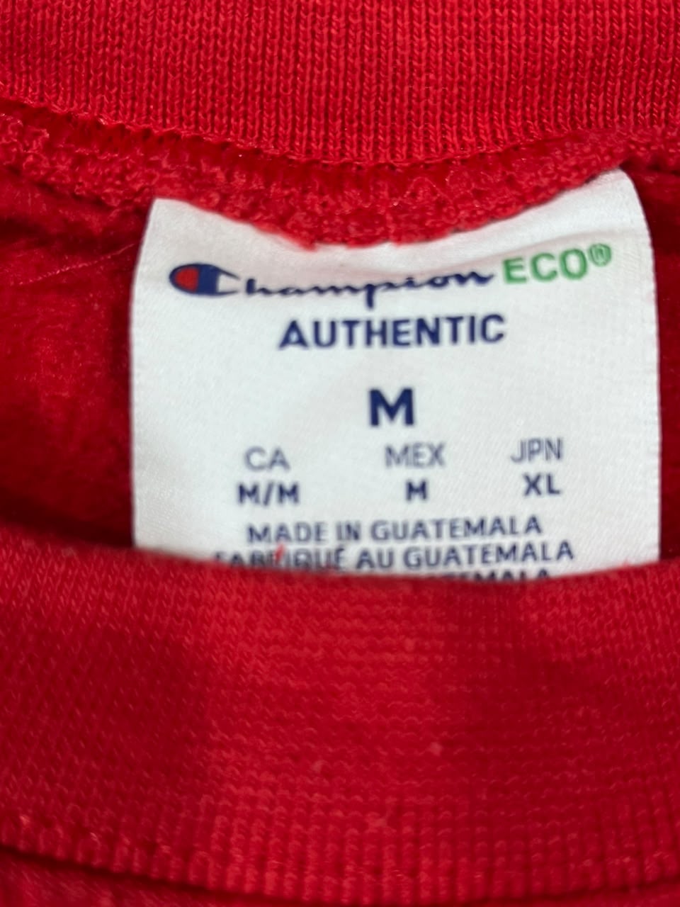 Close-up of a red garment's inner neck label showing Champion Authentic ECO branding and size M details.