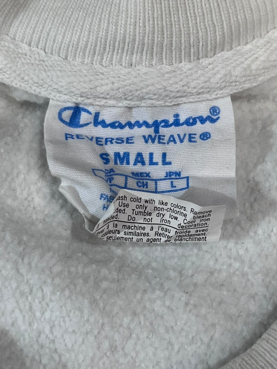 Close-up of a white Champion Reverse Weave sweatshirt tag labeled SMALL, attached to a white ribbed knit garment.