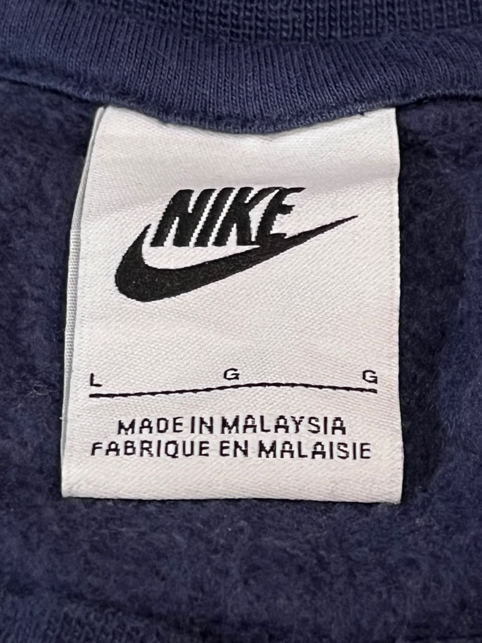 Close-up of a Nike garment label sewn onto a dark navy blue ribbed knit fabric, showing the black Nike swoosh logo and size information.