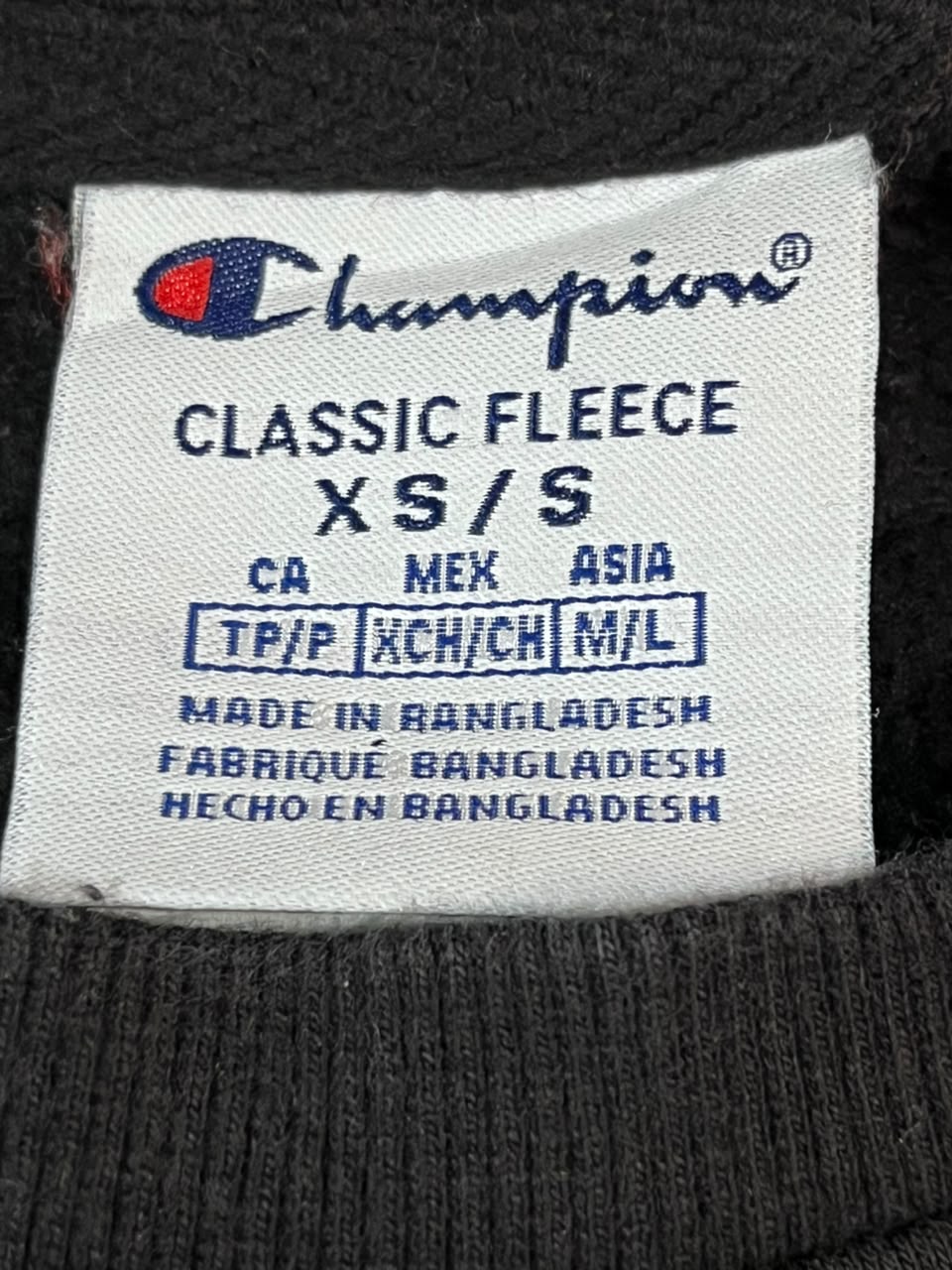 Close-up of a white Champion Classic Fleece XS/S garment label sewn onto a dark charcoal fleece fabric.