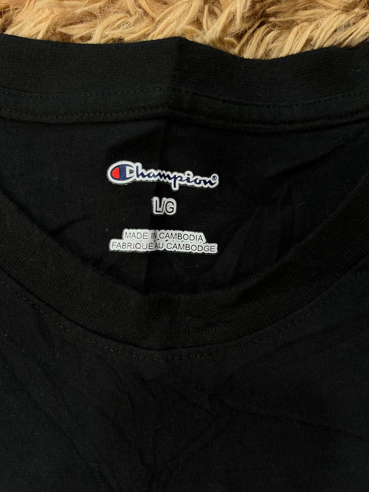 Close-up of a black Champion t-shirt neck label, showing the embroidered Champion logo and size information, resting on a textured tan surface.