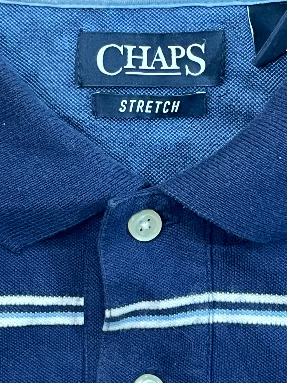 Close-up of a blue Chaps Stretch polo shirt collar, showing the brand label and white buttons.