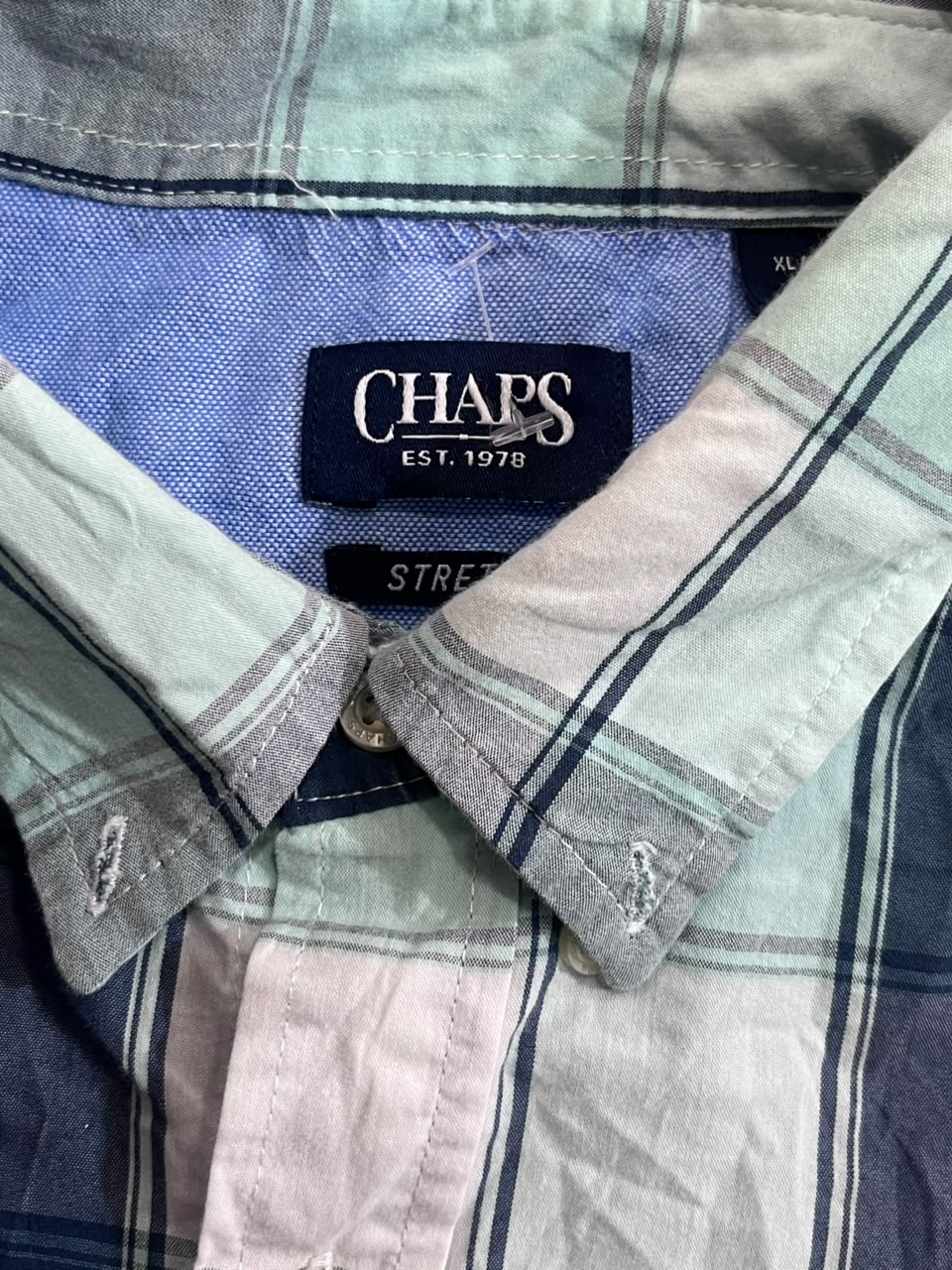 Close-up of a Chaps plaid button-down shirt collar, showing the brand label 'CHAPS EST. 1978' and the striped pattern in muted blue, gray, and pale pink tones.