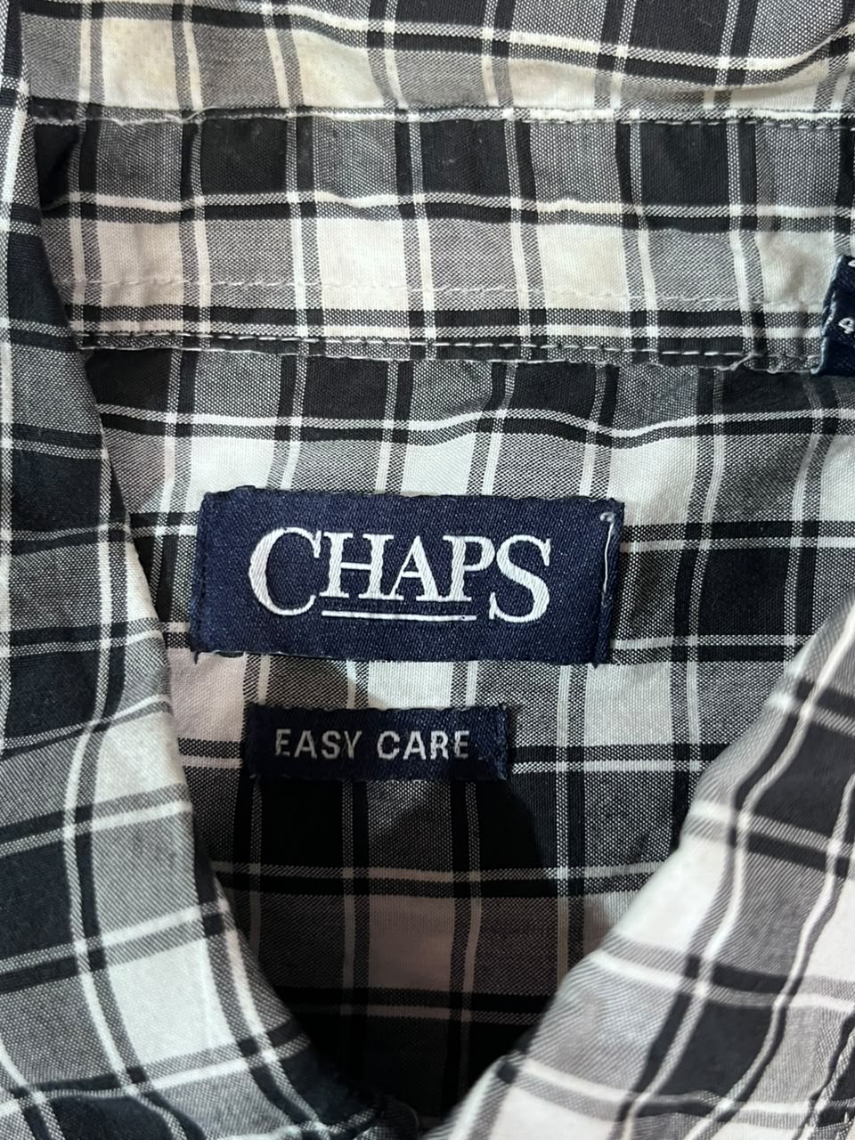 Close-up of a plaid shirt collar with a visible CHAPS brand label and an EASY CARE tag, showing a black, white, and gray pattern.