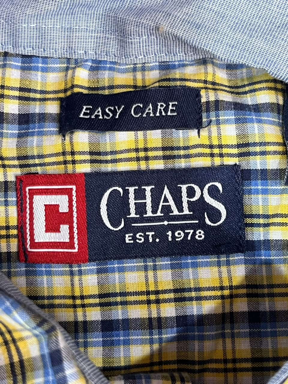 Close-up of a plaid shirt collar showing the brand label 'CHAPS EST. 1978' and an 'EASY CARE' tag on a yellow, blue, and white plaid fabric.