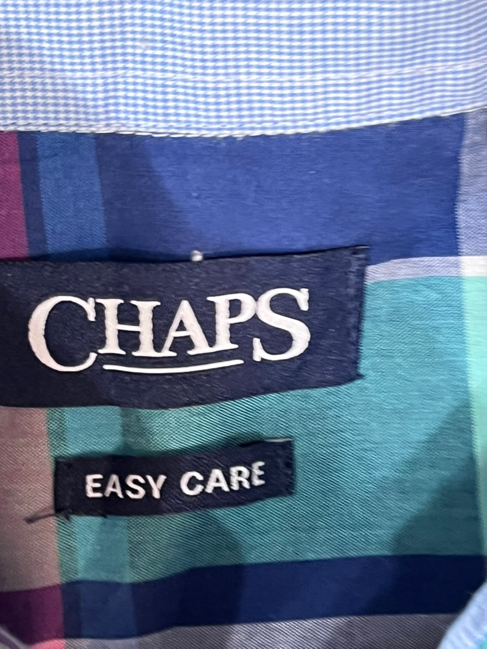 Close-up of a Chaps plaid shirt label showing the brand name 'CHAPS' and 'EASY CARE' on a blue, teal, and navy plaid fabric.